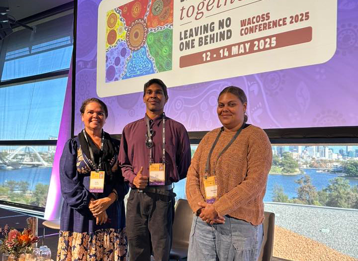 Three people stand smiling in front of a conference sign that reads, "Leaving No One Behind, WACOSS Conference 2025, 12-14 May." The mood is positive and inclusive.