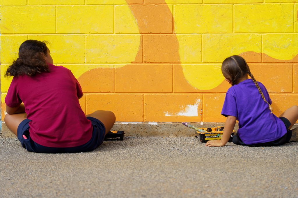 Two young girls sitting on the ground and painting a colourful wall