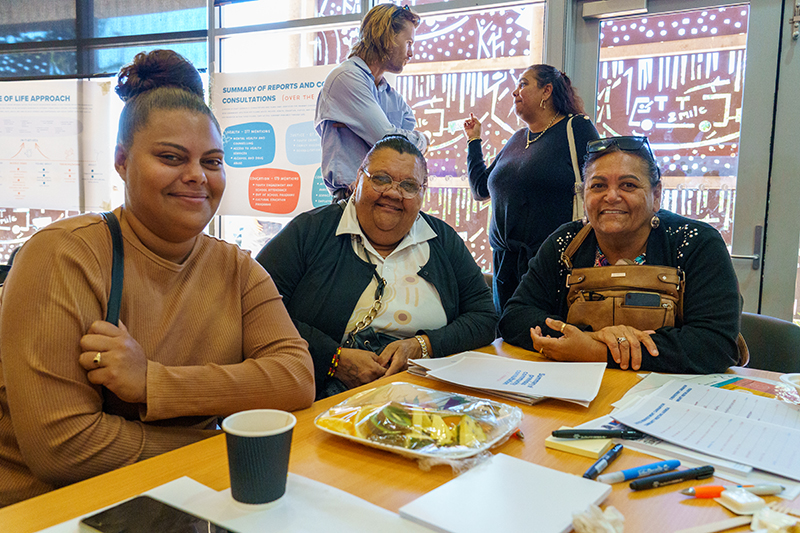 Three women smiling at the camera at a workshop for Carnarvon Common Ground. Two people are having a conversation in the background.