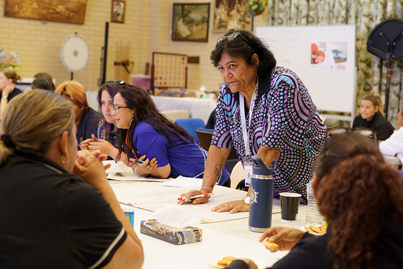 A woman leaning over the table with a pen and paper while talking to other women at a community workshop in Carnarvon