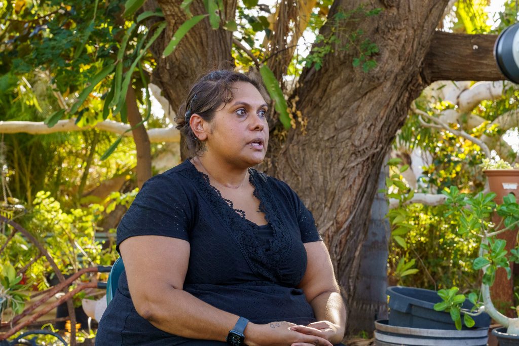 A woman sitting down and talking to a camera, in front of lush greenery and trees